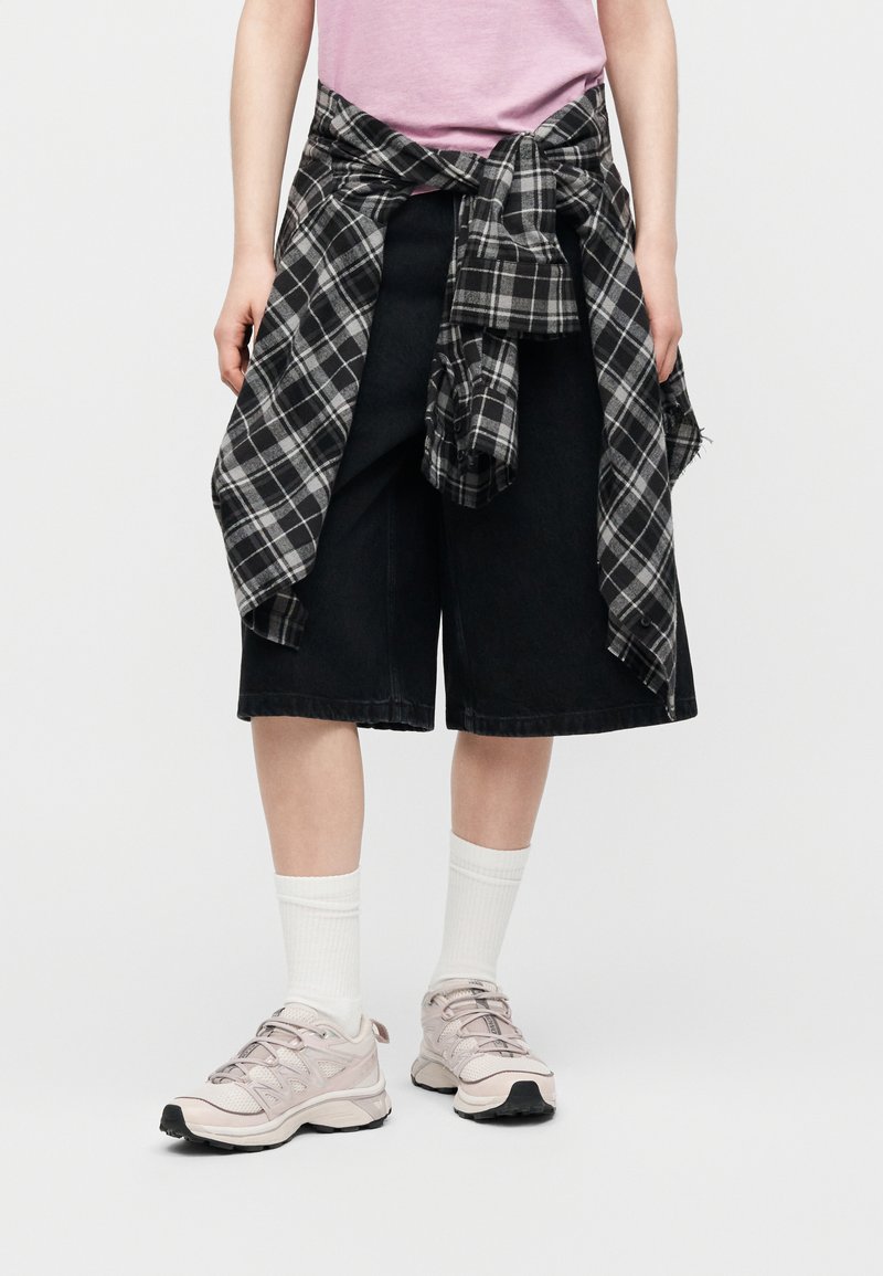 Person wearing black wide-leg shorts, a pink shirt, white socks, beige sneakers, with a black-and-white plaid shirt tied around the waist.