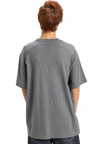 Personne avec des cheveux roux courts portant un T-shirt gris ample et un pantalon sombre, se tenant de dos contre un fond blanc.