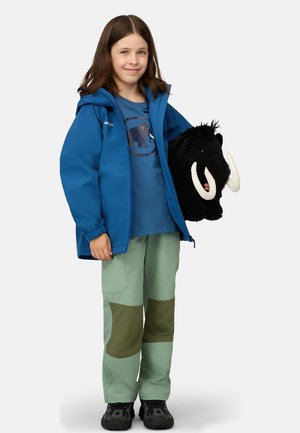 Ung flicka i blå jacka och gröna byxor håller ett stort svart plyschmammuthuvud med vita betar och ler mot kameran.