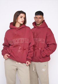 Red hoodies with "MULTIPLY" in white studs and graphics, paired with beige sweatpants featuring embroidered accents. Loose fit and soft texture.