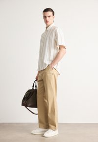 Korte mouwen, gekleed overhemd in crème met verticale strepen, gecombineerd met beige broek. Witte sneakers en een donkere tas maken de outfit compleet.