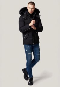 Czarna kurtka puchowa z kapturem z sztucznego futra, z kieszeniami na zamek, zestawiona z przetartymi niebieskimi dżinsami i czarnymi masywnymi botkami.