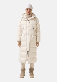 Lange gewatteerde jas in lichtcrème, met een capuchon, twee voorzakken en ritssluitingen. Gecombineerd met beige hoge laarzen en crèmekleurige broek.