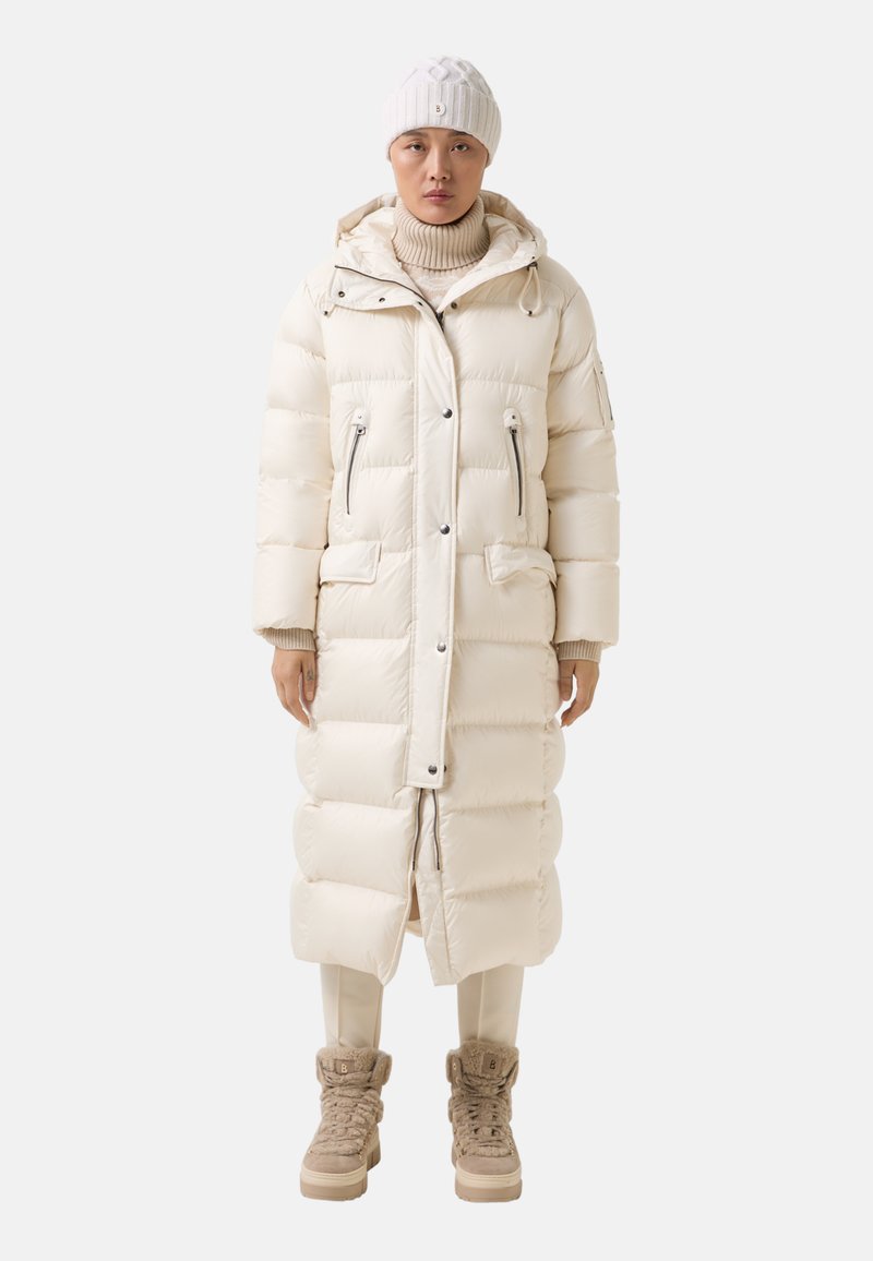 Lange gewatteerde jas in lichtcrème, met een capuchon, twee voorzakken en ritssluitingen. Gecombineerd met beige hoge laarzen en crèmekleurige broek.