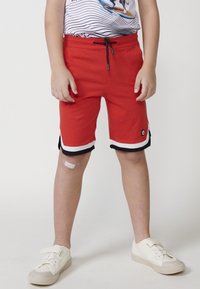 Pantaloni corti rossi in tessuto leggero, dotati di vita regolabile con cordino, inserti a strisce bianche e nere all'orlo e una patch con logo.
