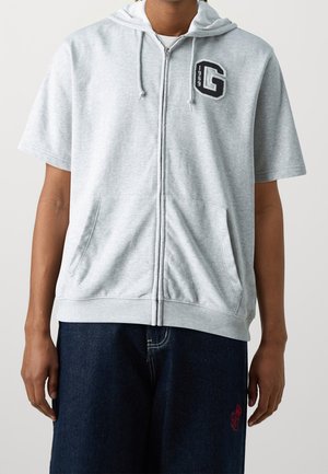 Sudadera con cremallera de color gris claro y mangas cortas, que presenta un logo en la parte frontal, dos bolsillos laterales y una capucha con cordón. Textura de tela suave.