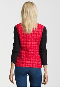 Czerwony ocieplany kamizelka z wzorem w jodełkę, pikowaną strukturą i stójką. Nosi na sobie czarną bluzkę z długim rękawem oraz niebieskie dżinsy.