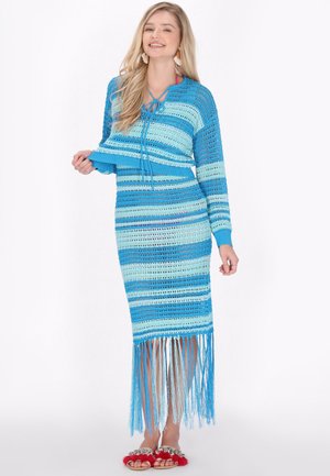 Frau lächelt, trägt ein blau gestreiftes Strickkleid mit langen Fransen am Saum und passende Hausschuhe mit roten Pelzakzenten.