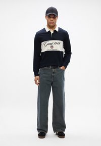 Polo cu mâneci lungi de culoare navy, cu guler crem și dungă pe piept, având inscripția 'Gant 1949'. Este purtat cu blugi lejeri, de culoare albastru deschis, și o șapcă neagră.