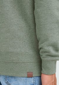 Sudadera verde con textura acanalada y mangas largas. Presenta un parche de logo de cuero marrón en la parte inferior izquierda. Combinada con jeans de mezclilla azul claro.