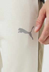 Nahaufnahme von hellbeige Hosen mit einem grauen Puma-Logo in der Hüftgegend und einer Hand, die neben den Hosen ruht.