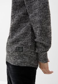 Suéter gris de punto con un patrón texturizado, mangas largas y una etiqueta en el dobladillo inferior. La tela parece suave y cálida.