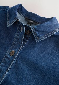 Col de chemise en denim avec les deux premiers boutons fermés, étiquette de marque "NEXT" visible et étiquette "Fabriqué au Bangladesh" sur un fond clair.