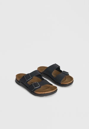 Paar schwarze Sandalen mit doppelten Riemen und verstellbaren Schnallen, braun gepolsterte Innensohle und schwarze Sohle auf hellem Hintergrund.