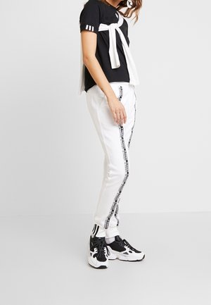 Kvinde iført sort t-shirt, hvid sweatshirt bundet over skuldrene, hvide Adidas træningsbukser med sorte logo-striber, og sort-hvide sneakers.