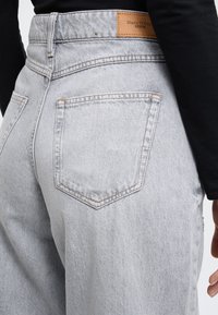 Világosszürke, magas derekú farmernadrág hátul zsebekkel és barna bőr címkével. Sima textúra, látható varrási részletekkel.