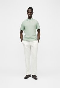 Homme debout, les mains dans les poches, portant un polo à manches courtes vert clair, un pantalon blanc et des chaussures habillées marron foncé sur fond blanc.