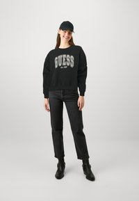 Felpa nera in cotone con logo "GUESS" in bianco e grigio, abbinata a jeans neri e stivali alla caviglia. Il modello indossa un berretto nero.