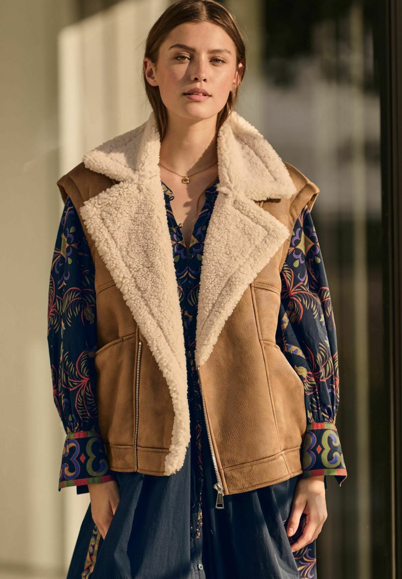 Tan faux-shearling-vestti, jossa on leveä kaulus, vetoketjusulku ja sivutaskut, päällä sininen kuvioitu mekko, jossa on värikkäät hihansuut.