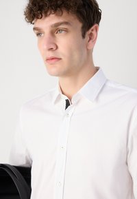 Camisa blanca de botones con un cuello clásico, que presenta una textura suave y un detalle de acento negro en el cuello. El modelo sostiene un bolso negro.