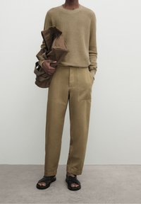 Ulltröja i beige, håller i en brun väska. Bruna byxor med vida ben och fickor. Svarta sandaler med platt sula. Neutral bakgrund.