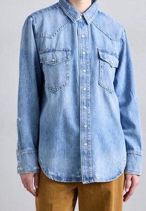 Personne portant une chemise en denim bleu clair à boutons avec deux poches poitrine et un pantalon en velours côtelé marron, devant un fond clair uni.