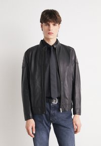 Black leather jacket with a stand-up collar, zip closure, and quilted shoulder accents. Paired with a black shirt and jeans.
