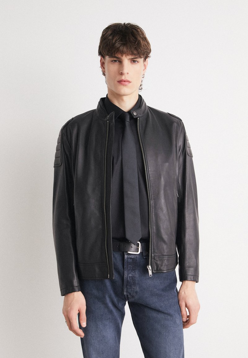 Black leather jacket with a stand-up collar, zip closure, and quilted shoulder accents. Paired with a black shirt and jeans.