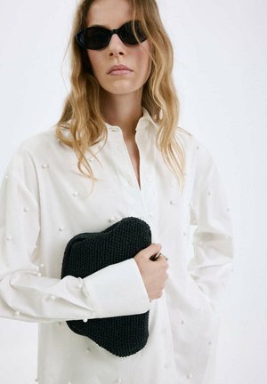 Žena má na sebe čierne úzke slnečné okuliare a bielu košeľu s malými štruktúrovanými bodkami, drží čiernu pletenú kabelku na bielom pozadí.