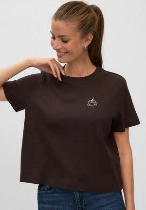 Femme souriante avec une queue de cheval portant un t-shirt marron foncé avec une petite broderie blanche de tasse de café sur le côté gauche de la poitrine.