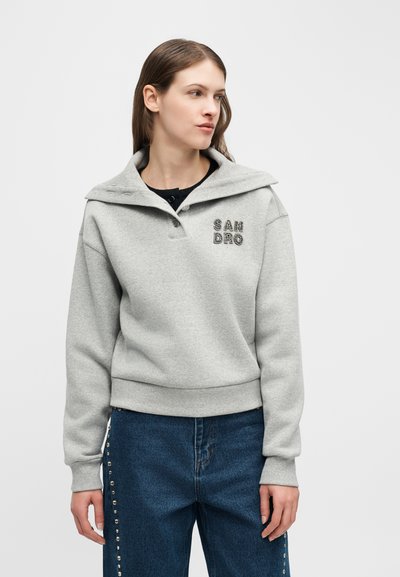 Νεαρή γυναίκα που φοράει ανοιχτό γκρι φούτερ με το κείμενο "SANDRO" και μπλε τζιν διακοσμημένα με ασημένια κρόσσια στα πλαϊνά, κοιτάζοντας προς τα αριστερά.