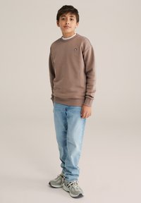 Fiú áll, barna pulóvert, világoskék farmert és szürke tornacipőt viselve, egyszínű bézs háttér előtt.