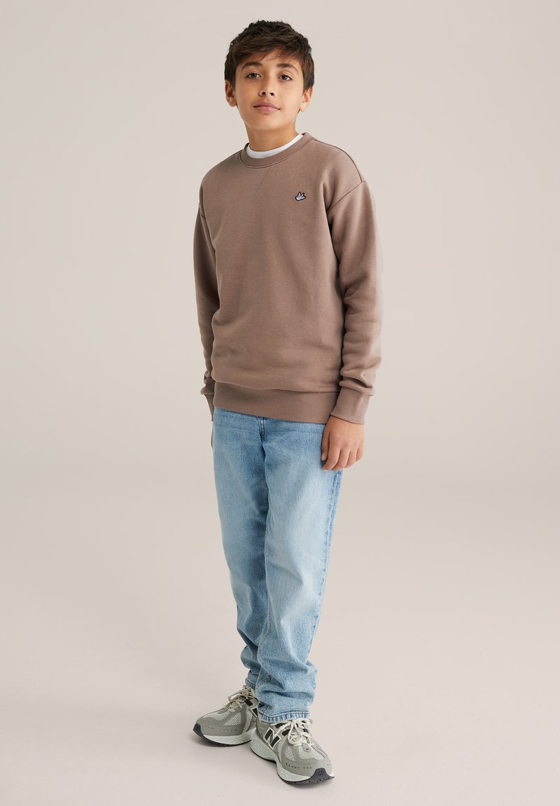 Fiú áll, barna pulóvert, világoskék farmert és szürke tornacipőt viselve, egyszínű bézs háttér előtt.