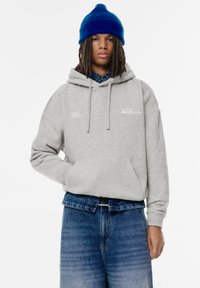 Grå huvtröja med framficka, snoddar och tryckta logotyper. Bärs med blå jeans och en blå stickad mössa.