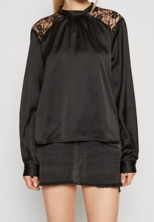 Kvinde iført en sort satinbluse med lange ærmer og blonder på skuldrene, kombineret med en sort denim mininederdel med rå kant.