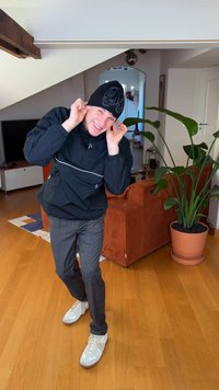 Coupe-vent noir avec bande blanche, pantalon à carreaux gris, baskets blanches et un bonnet noir. Environnement intérieur avec une plante verte en pot.