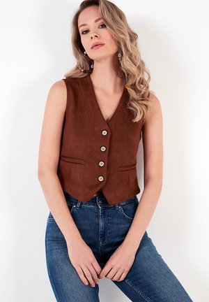 Brun ruskind cropped vest med V-udskæring, fem knaplukninger foran og to sidelommer, parret med blå denimjeans.