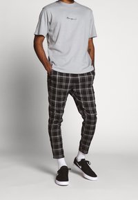Man in een lichtgrijze T-shirt, zwart-wit geruite broek, witte sokken en zwarte Nike-sneakers, staand met de handen in de zakken.