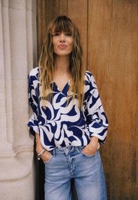 Marineblaue und weiße Bluse mit V-Ausschnitt und lockeren Ärmeln, kombiniert mit hellblauen, hoch taillierten Jeans. Holzhintergrund.