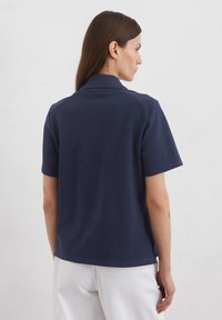 Navy Poloshirt aus Baumwolle, mit kurzen Ärmeln, Kragen und geradem Saum. Es gibt seitliche Schlitze für Bewegungsfreiheit.