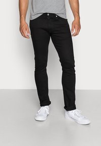 Person med slim-fit svarta jeans, ljusgrå t-shirt och vita sneakers som står på en ljusgrå golv mot en vit vägg.