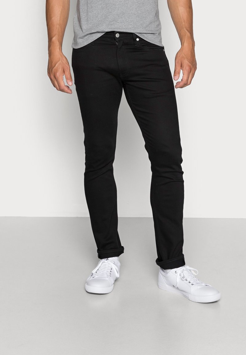 Person med slim-fit svarta jeans, ljusgrå t-shirt och vita sneakers som står på en ljusgrå golv mot en vit vägg.