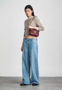 Hellbrauner Strickcardigan mit rundem Halsausschnitt, kombiniert mit weiten blauen Jeans aus Denim mit weißen Punkten. Ergänzt durch eine bordeauxfarbene gesteppte Handtasche.