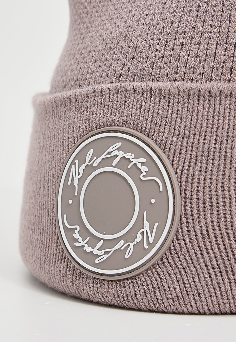 Beige gebreide beanie muts met een gestructureerd oppervlak en een circulaire rubberen patch met witte tekst en een logo aan de voorkant.