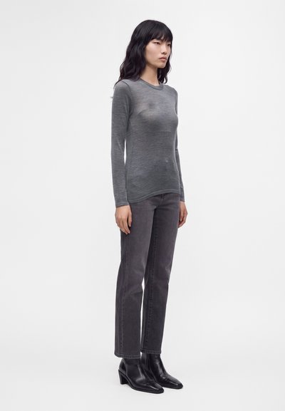 Femme debout portant un haut gris ajusté à manches longues, un jean droit gris foncé et des bottines noires, sur un fond clair uni.