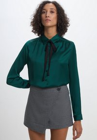 Blusa di raso verde smeraldo con un fiocco nero, dotata di colletto e chiusura a bottoni, abbinata a una gonna avvolgente in pattern scozzese bianco e nero.