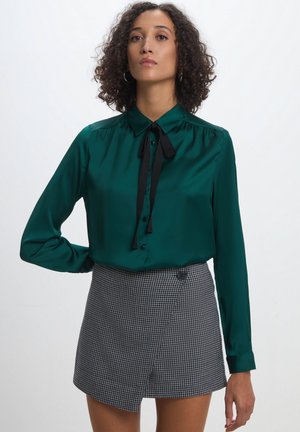 Blusa di raso verde smeraldo con un fiocco nero, dotata di colletto e chiusura a bottoni, abbinata a una gonna avvolgente in pattern scozzese bianco e nero.