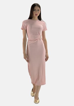 Model trägt ein enges rosa Kleid mit einer Seiten-Schnalle, kombiniert mit goldenen Schuhen, vor einem schlichten weißen Hintergrund.