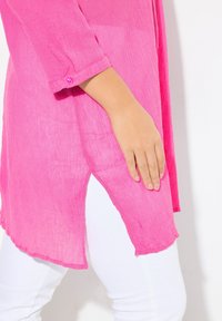 Roze tuniek met een gerimpelde textuur, driekwartmouwen en een zijsplit, gecombineerd met witte broek. Eenvoudige knoopdetails op de mouw.