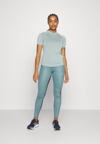 Kvinne iført lys blå treningsskjorte og tights med mørkeblå joggesko, stående mot en ensfarget hvit bakgrunn.